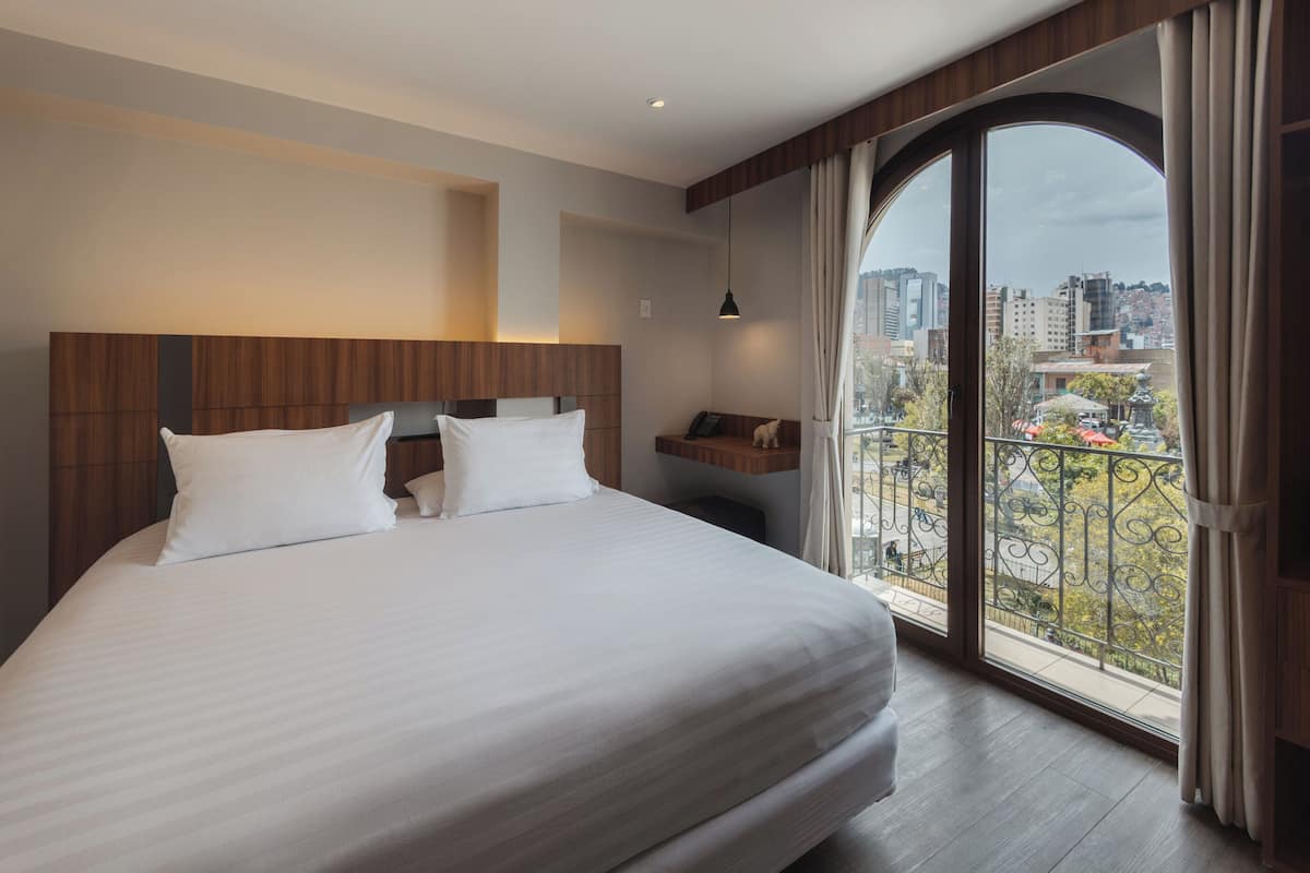 Une chambre d’hôtel avec un grand lit, une tête de lit en bois, une table de chevet avec une lampe et une vue sur la ville par une grande fenêtre.