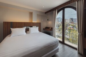 Habitación de hotel con una cama grande, cabecera de madera, mesita de noche con lámpara y vistas a la ciudad a través de un ventanal amplio.