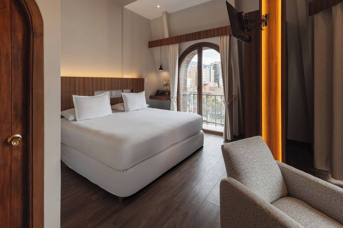 Une chambre d’hôtel avec un grand lit, une tête de lit en bois, un téléviseur fixé au mur et un balcon avec des rideaux.