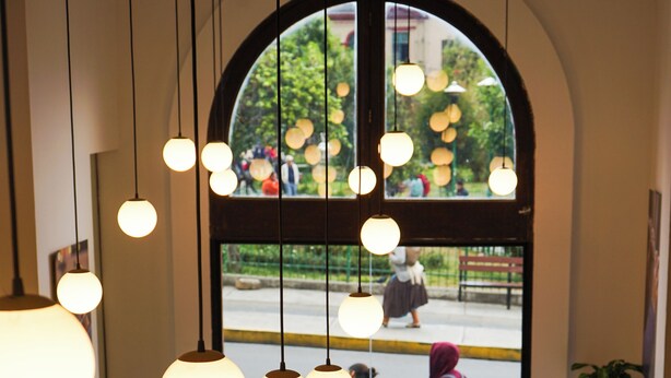 A restaurant with a long wooden table, black chairs, hanging spherical light fixtures, and a large arched window overlooking a street. - DREAM By Stannum Hotel (La Paz)