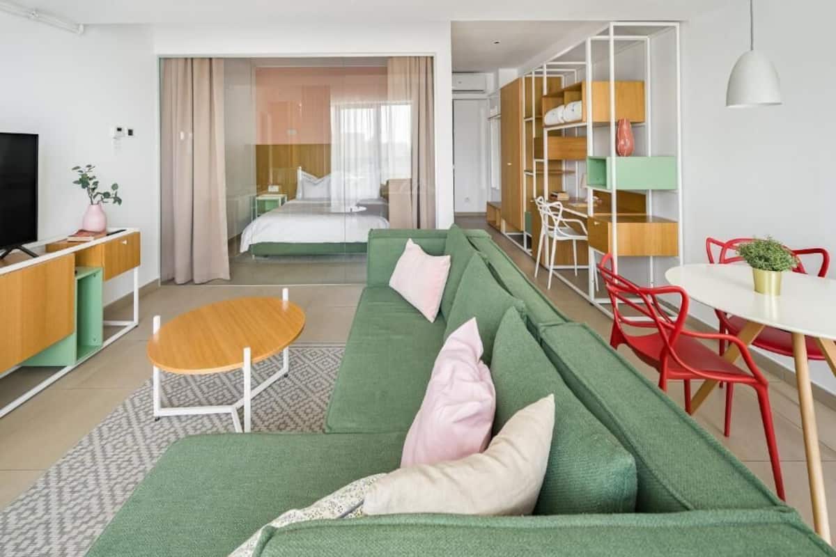 A modern hotel room with a green sofa, a wooden dining table, and a separate bedroom with a bed and curtains.