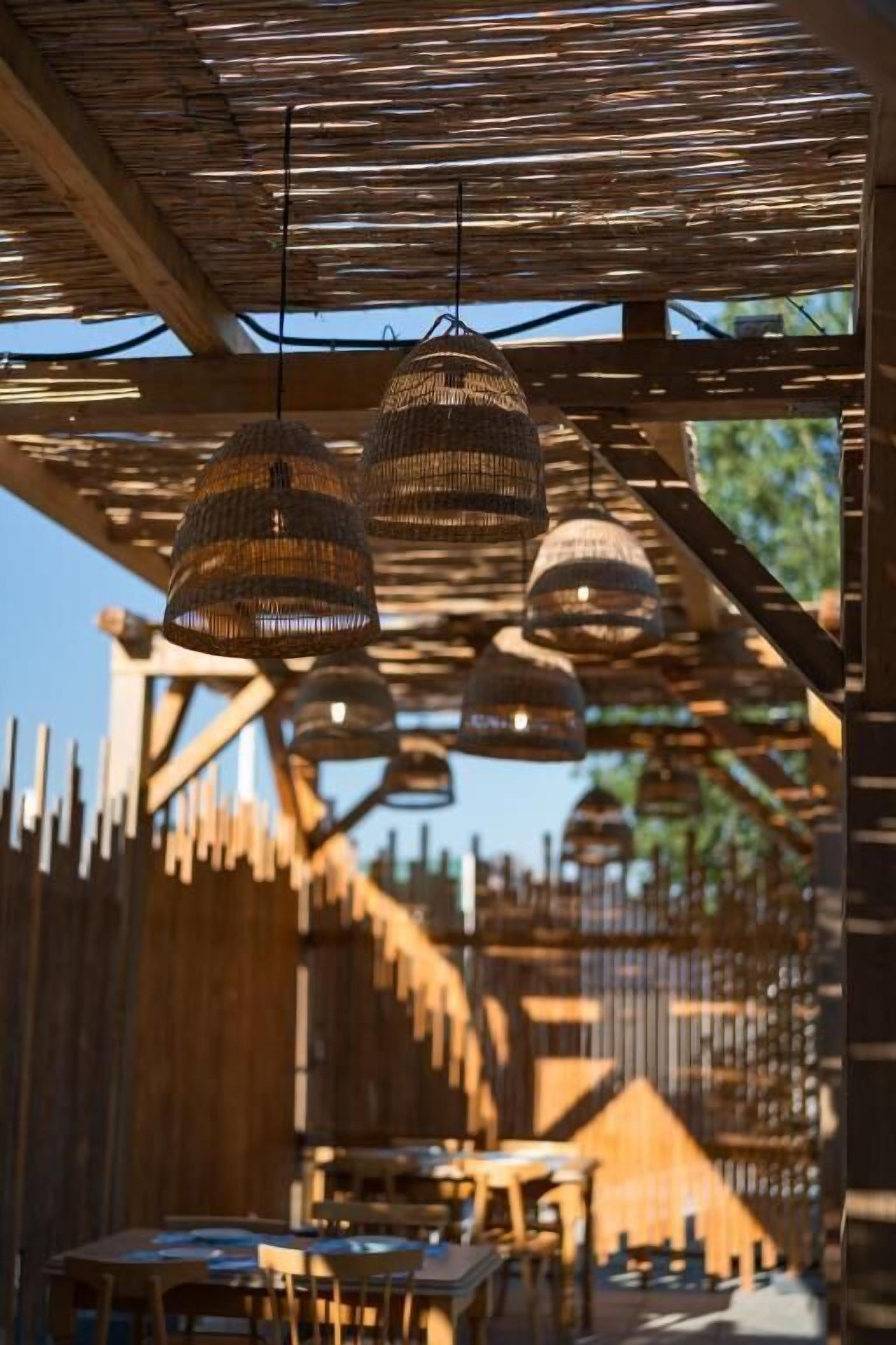Un área de comedor al aire libre con mesas y sillas de madera, y lámparas de mimbre colgantes.
