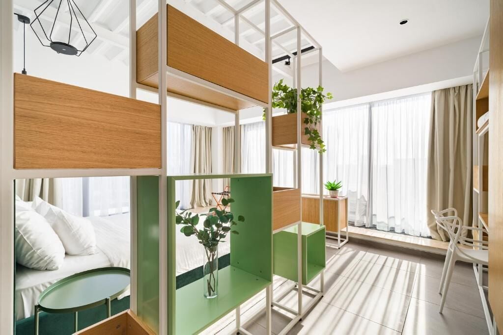 A modern hotel room with a bunk bed, a small desk with a plant, a chair, and a large window with curtains.