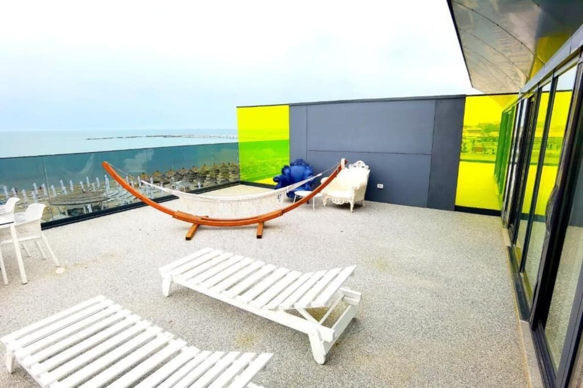 A balcony with white lounge chairs, a hammock, and a view of the sea.