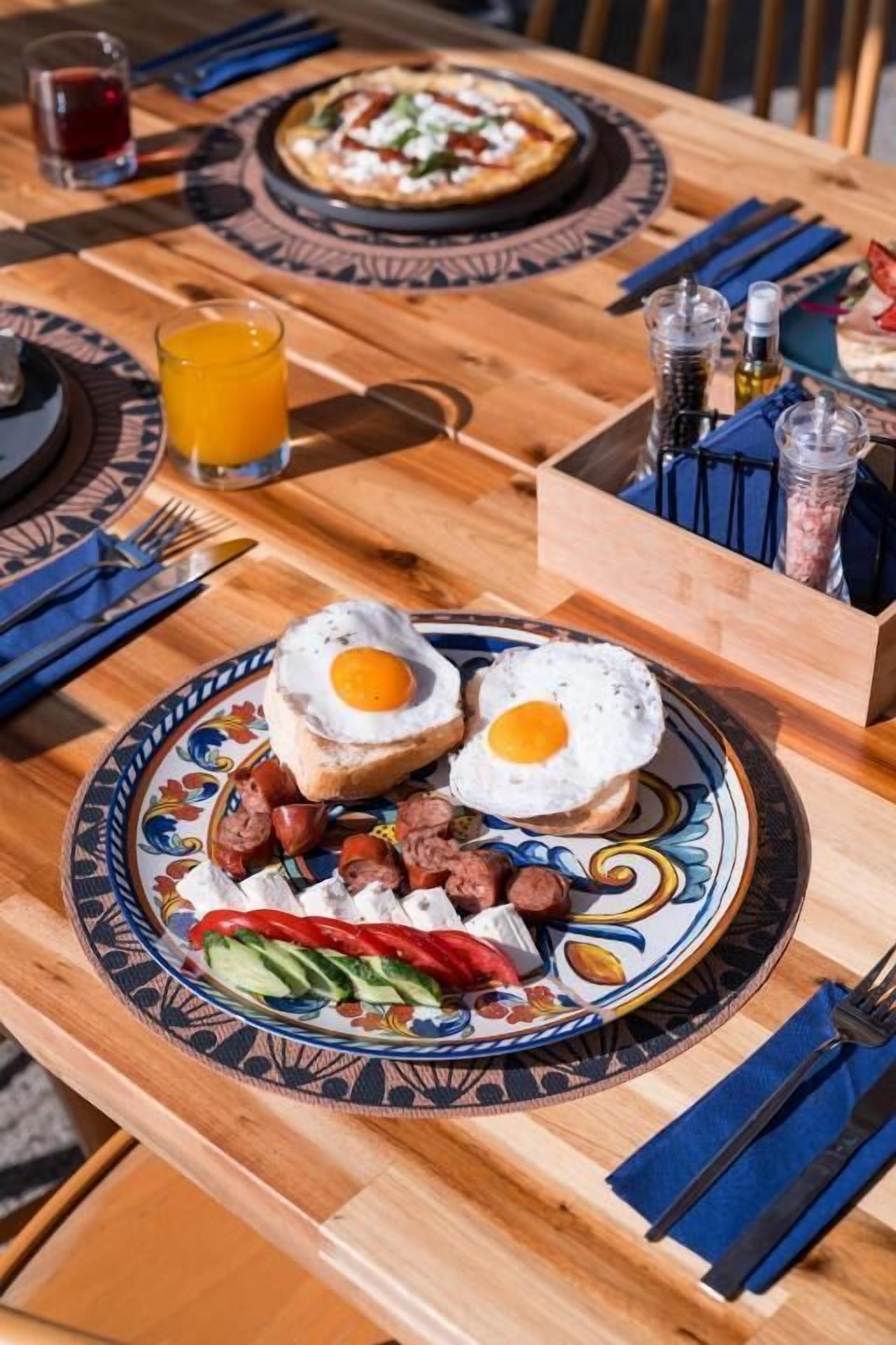 Una escena de desayuno con un plato de huevos, salchichas y verduras, un vaso de jugo de naranja y una pizza sobre una mesa de madera.