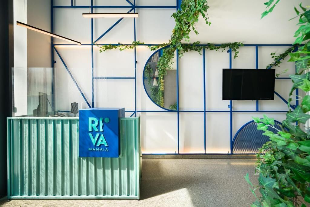 Un espace d’accueil avec une enseigne bleue portant l’inscription « RIVA MAMMA », un téléviseur fixé au mur et un mur agrémenté d’éléments structurels bleus et de plantes.
