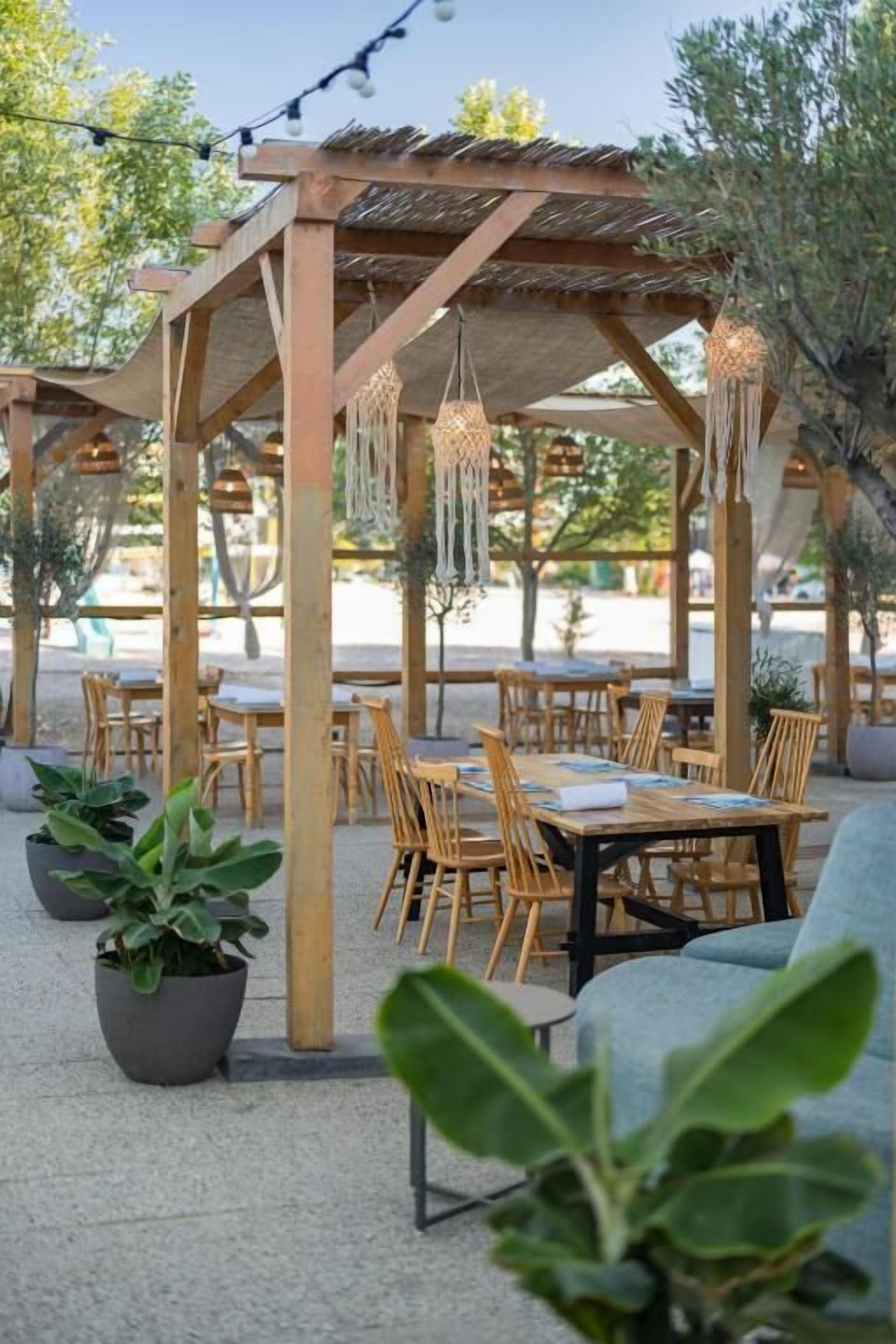 An outdoor seating area with wooden tables and chairs, hanging lights, and potted plants.
