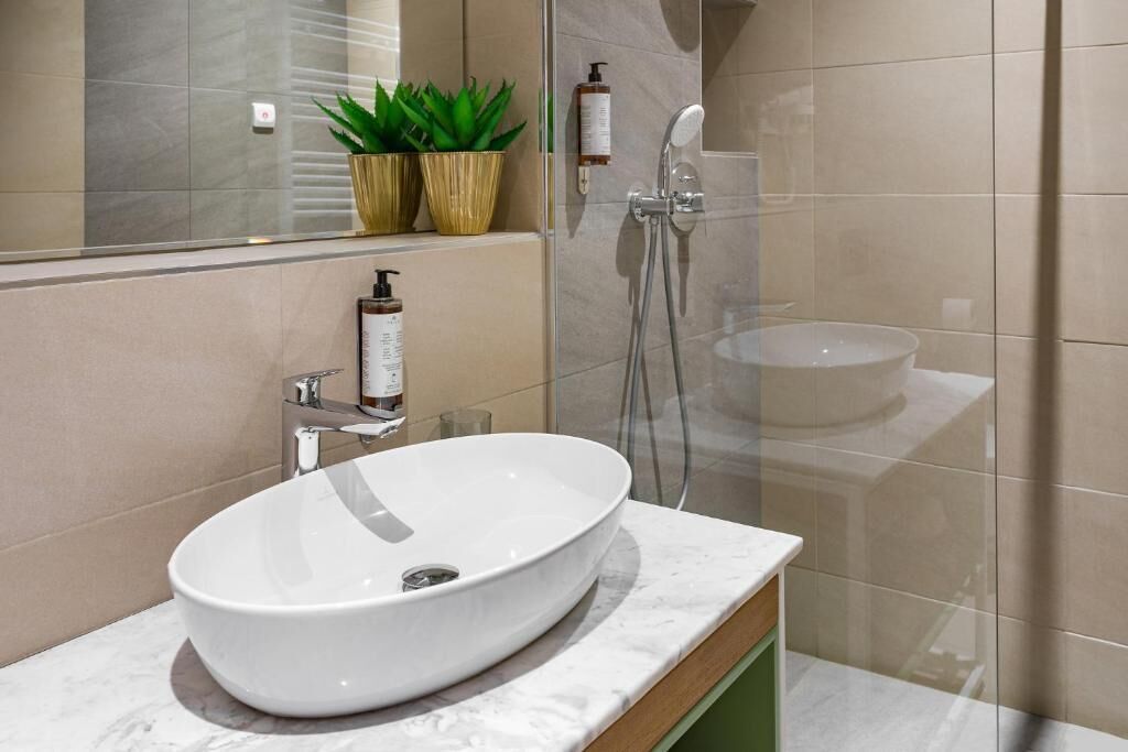 A modern bathroom with a large oval sink, a handheld showerhead, and two potted plants on the vanity.