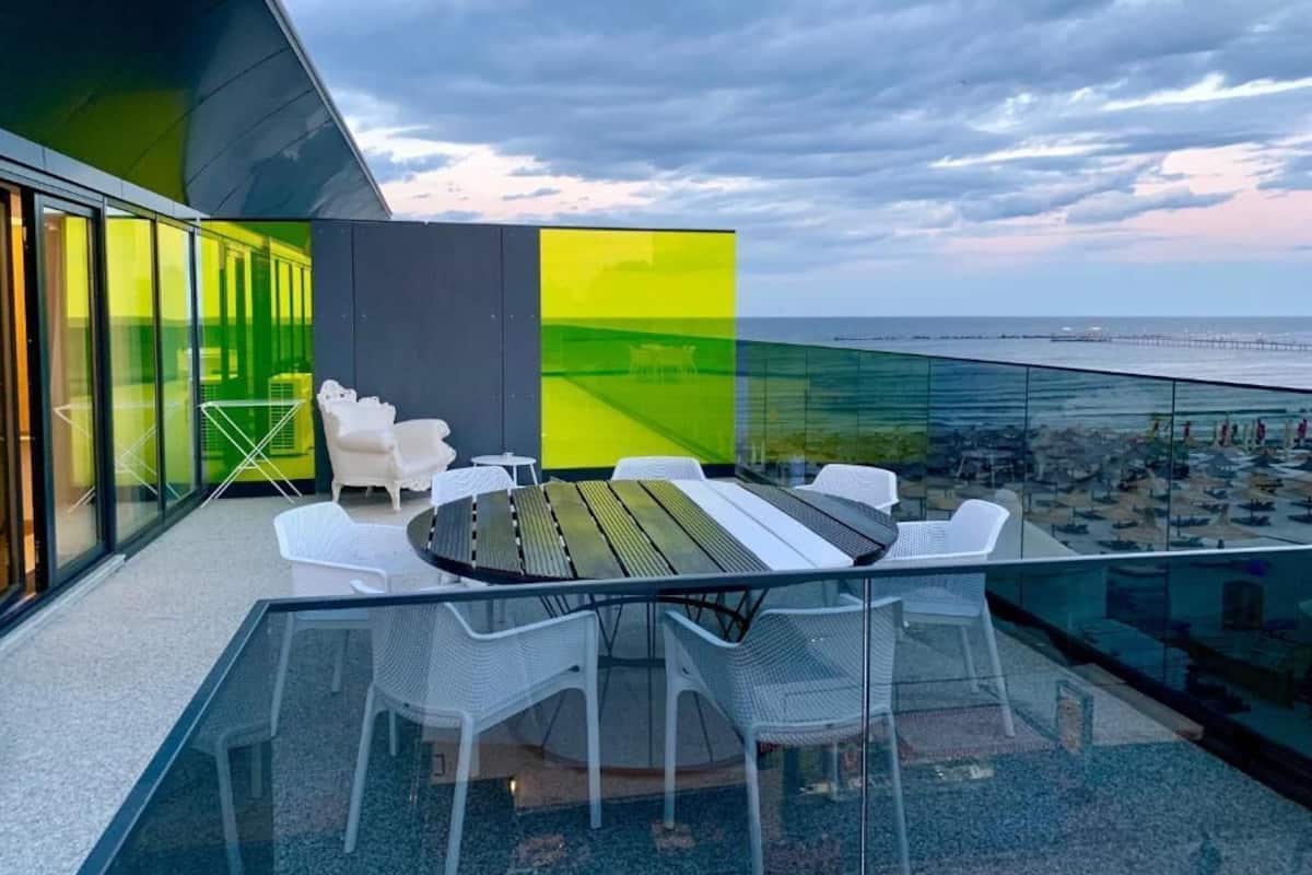 A modern outdoor dining area with a glass table and chairs, overlooking a beach and a pier.