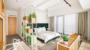 A modern hotel room with a bed, a desk, a chair, a green shelving unit, and a potted plant.