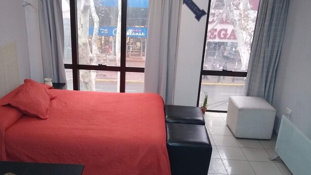 A hotel room with a bed, a small table, a bench, and a window with a view of the street.