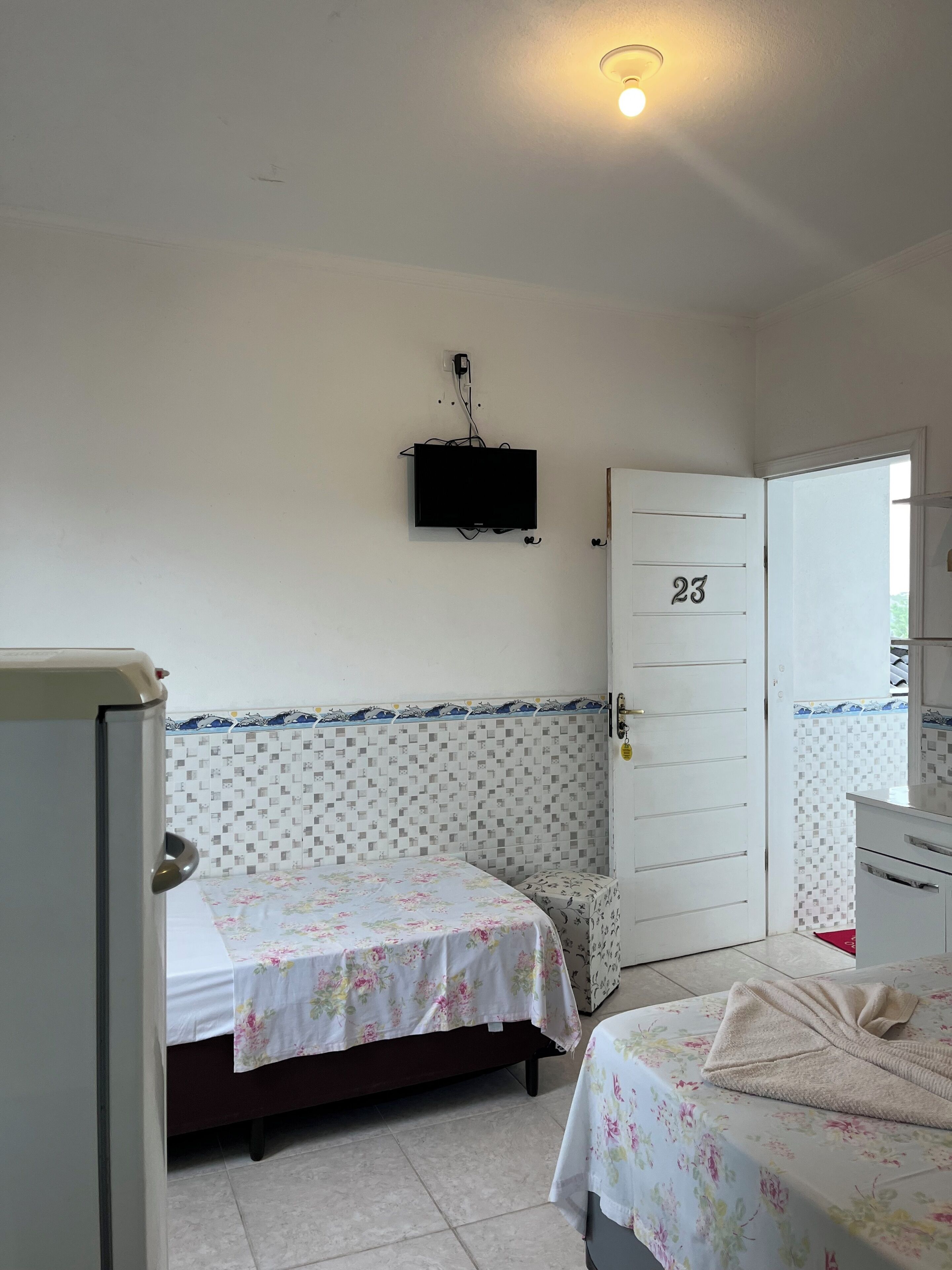 Quarto com duas camas, televisão fixada na parede, porta com o número 23 e parede revestida de azulejos.