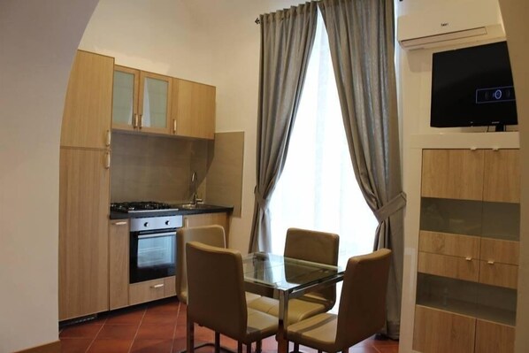 Una cocina compacta con estufa a gas, horno y armarios de madera. Hay una mesa de comedor con cuatro sillas y un televisor montado en la pared.