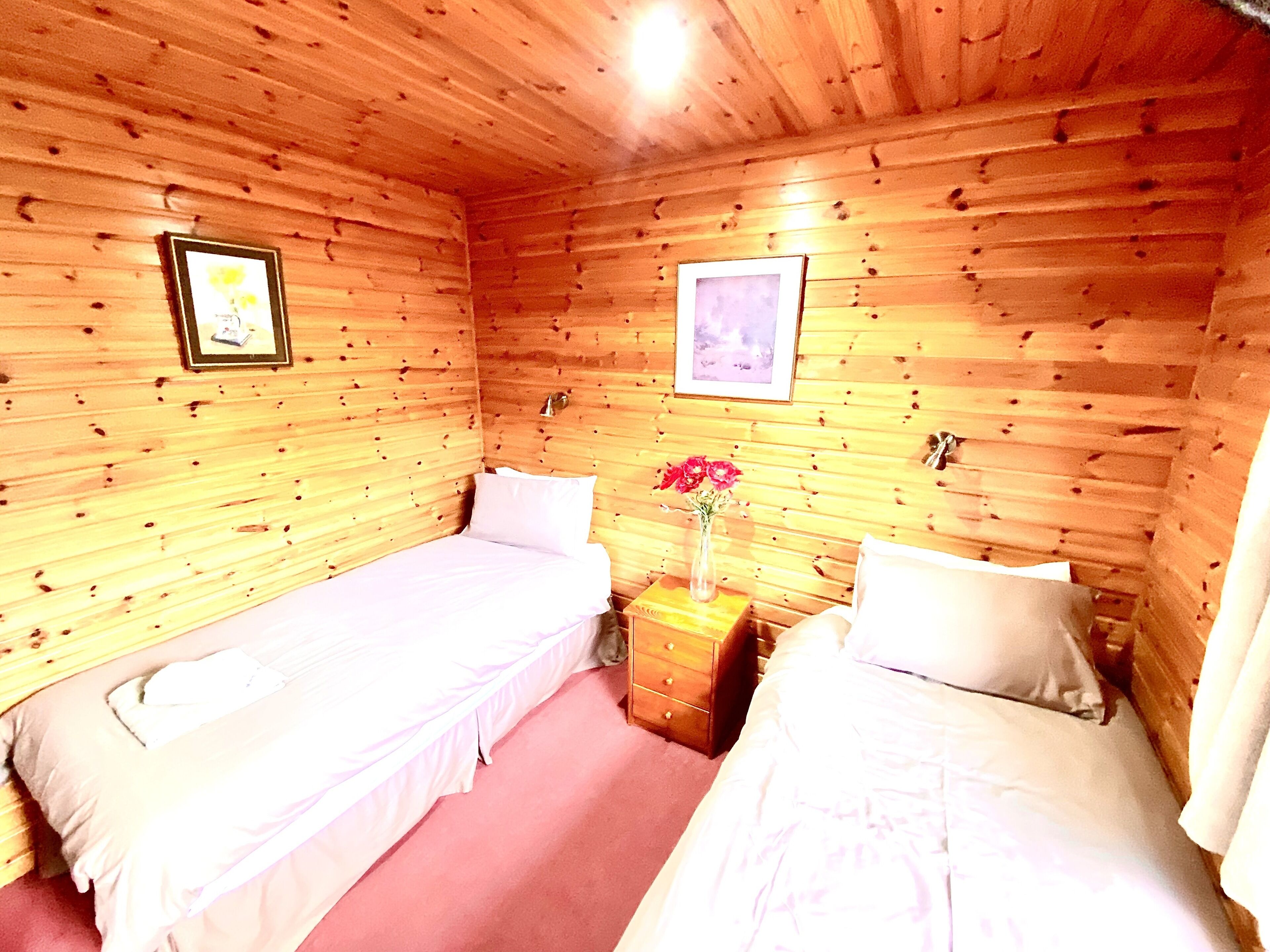 A cabin-style room with two single beds, a wooden ceiling, and a small wooden nightstand with a vase of flowers.