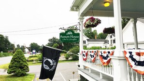 Un porche con un cartel de Sutton House Bed & Breakfast, banderas y macetas colgantes de flores.