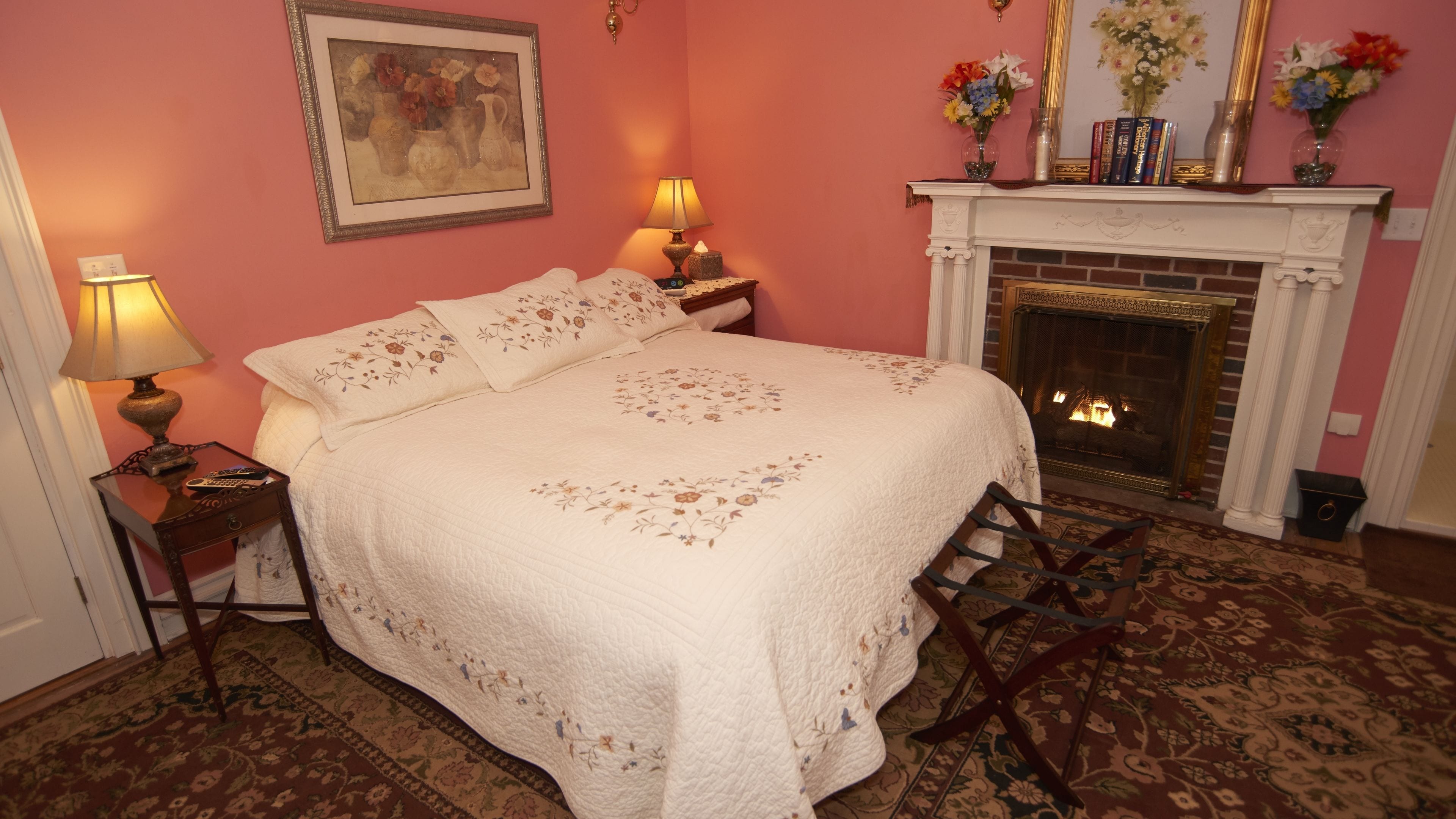 A bedroom with a large bed, two bedside tables with lamps, a fireplace, and a framed painting on the wall.