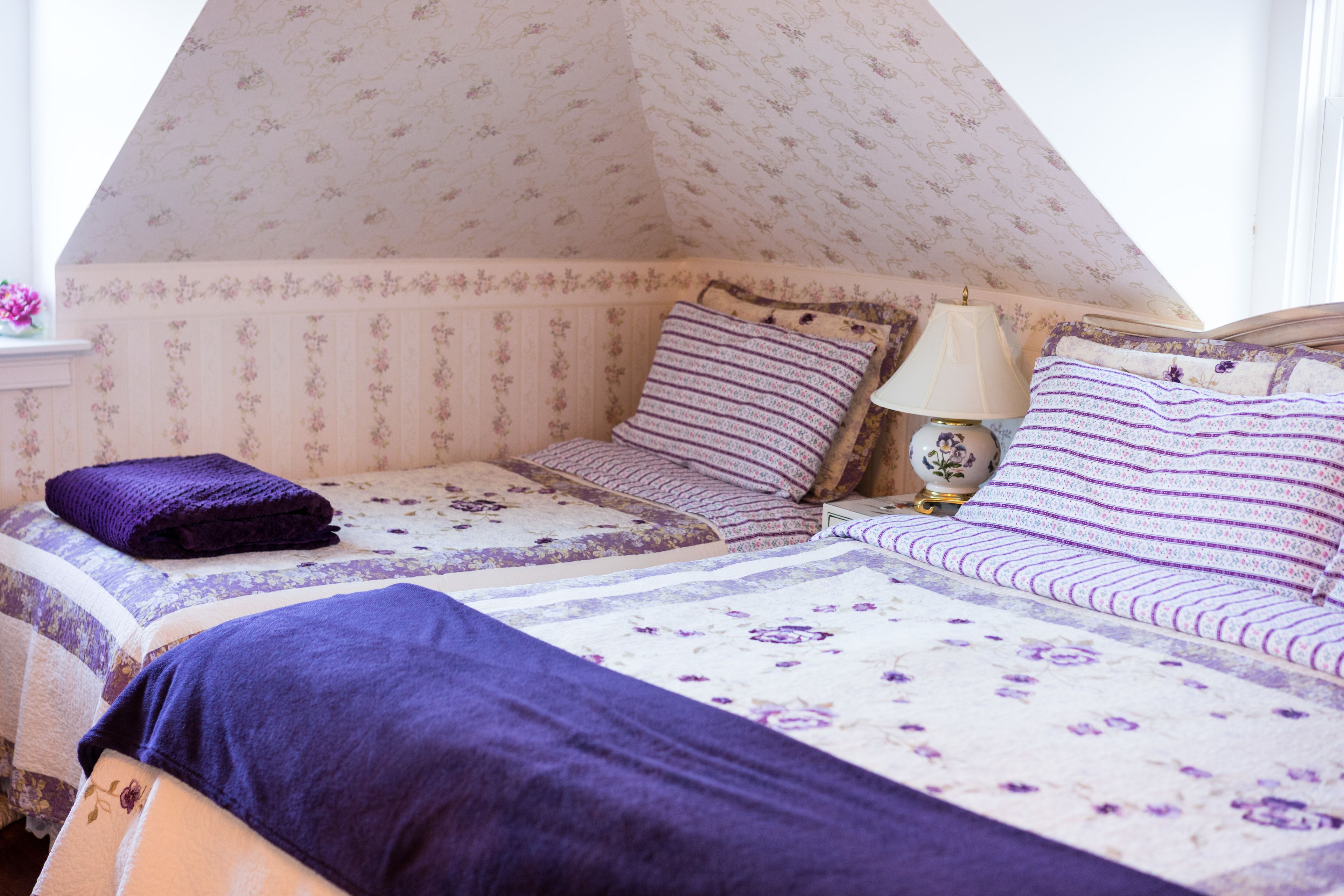 Un dormitorio con techo inclinado, papel tapiz floral y cama con ropa de cama morada.