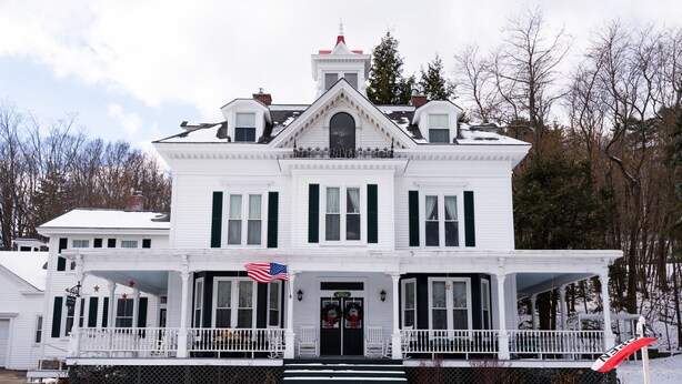 Una amplia casa blanca de dos pisos con un porche delantero, una bandera y un terreno cubierto de nieve.