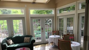 Una habitación iluminada por el sol con un sofá verde, dos mesas redondas con manteles blancos y un banco verde fuera de una puerta de cristal.