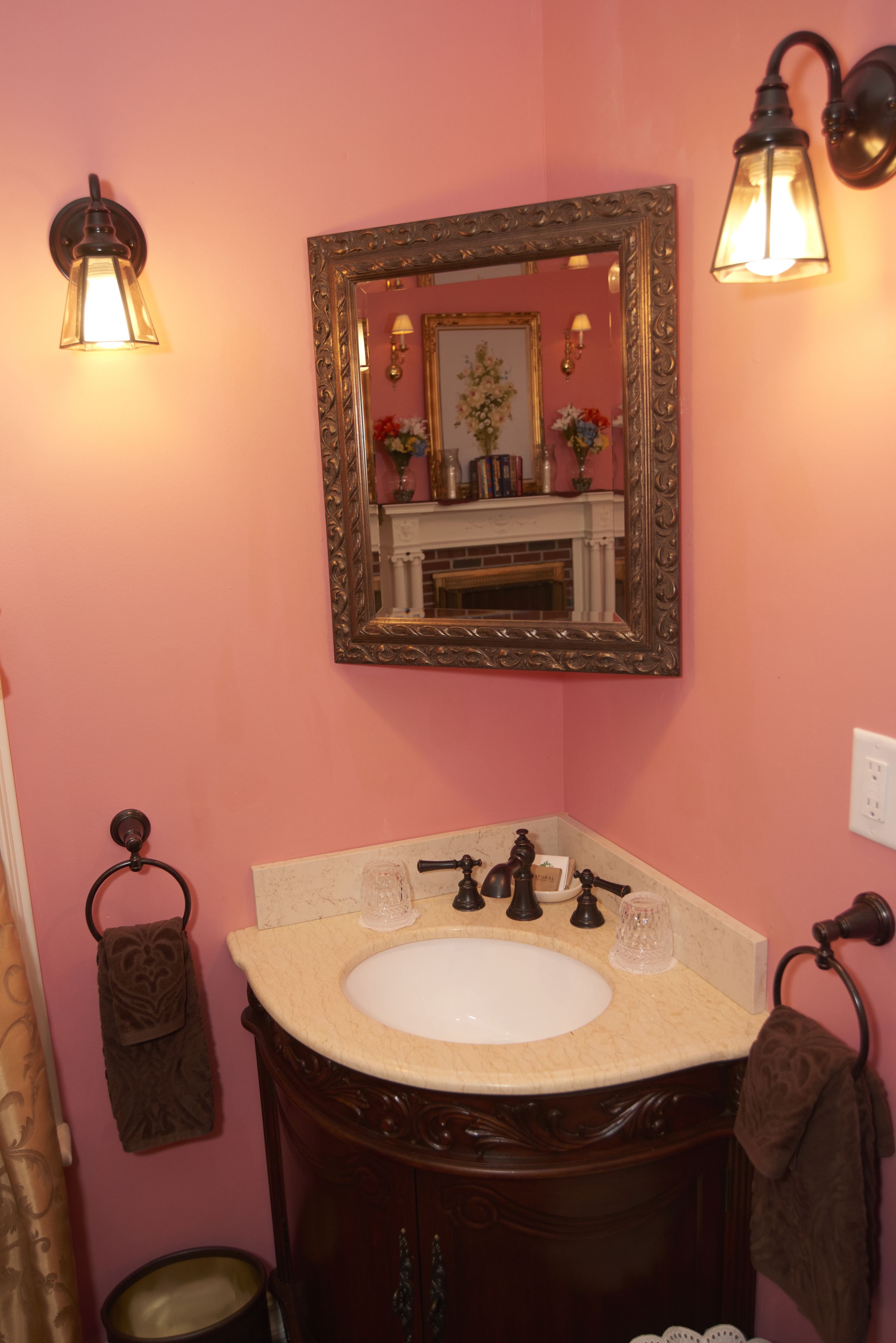 A bathroom with a pink wall, a wooden vanity, a white sink, two wall-mounted lights, a mirror with an ornate frame, and a towel rack with two towels.