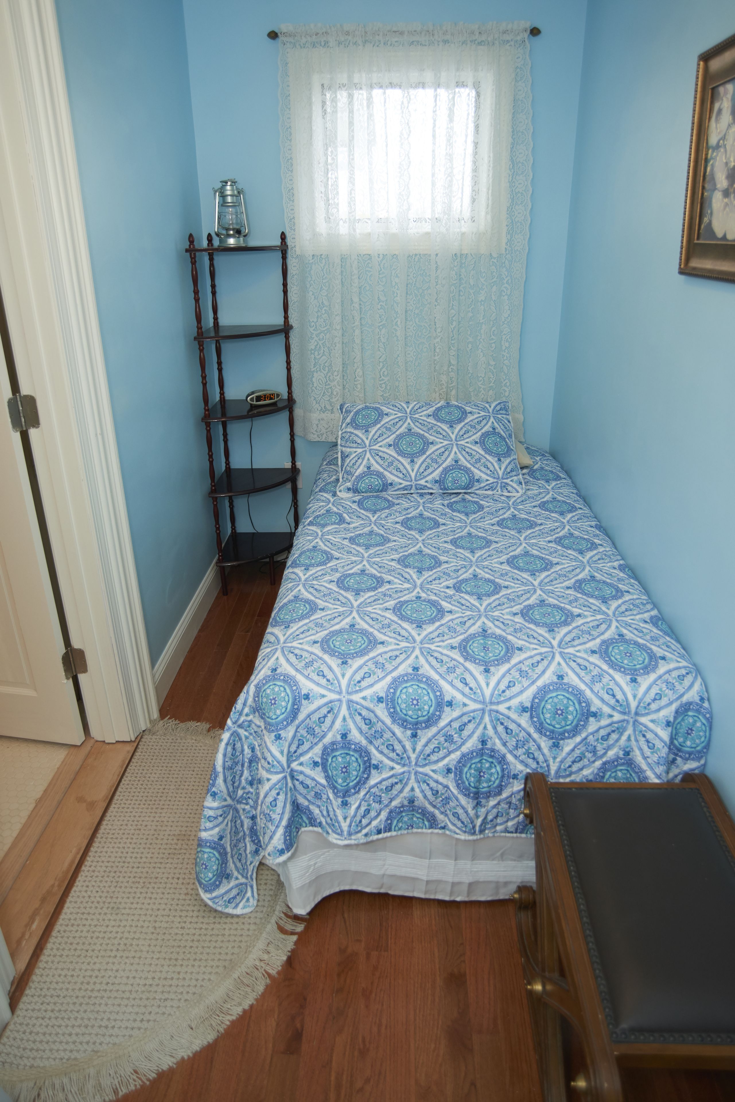 A bedroom with a bed, a wooden shelf, a small table, and a window with a sheer curtain.