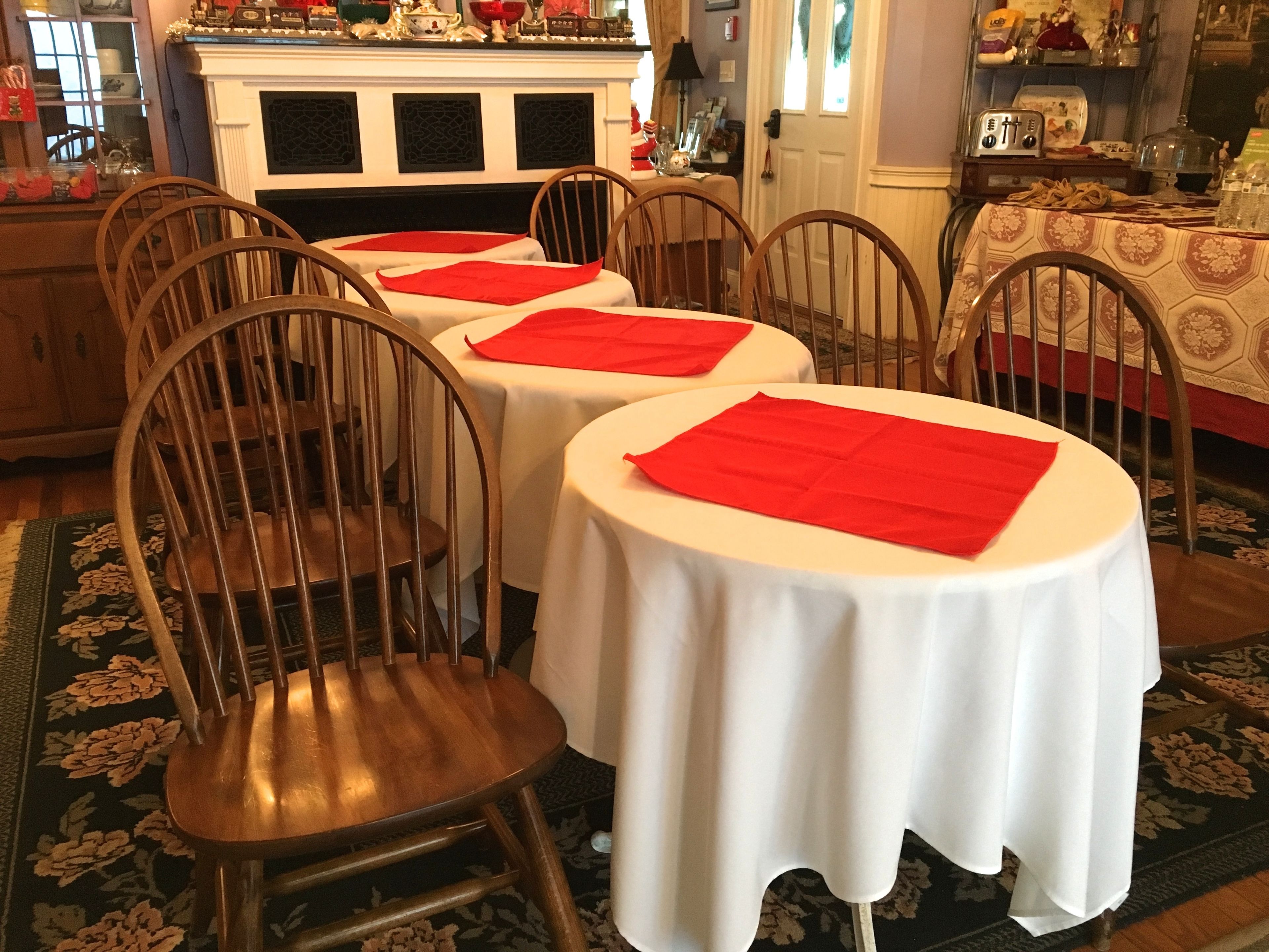 A dining area with two round tables, each with a white tablecloth and red placemats. The chairs are wooden with a curved backrest.