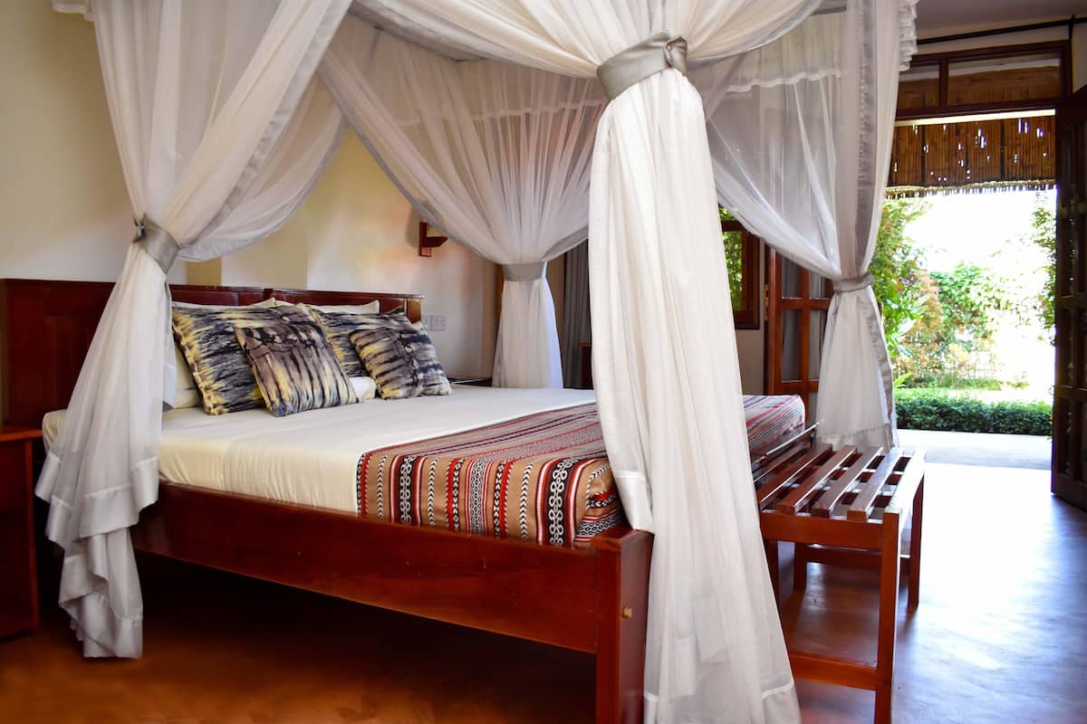 A four-poster canopy bed with a wooden frame and white bedding, a bench at the foot of the bed, and a view of greenery outside.