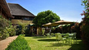 Een tuin met een gebouw met rieten dak, groene stoelen en een tafel onder een grote parasol.