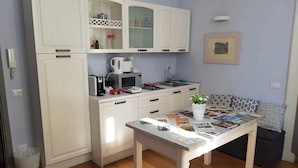 A compact kitchen with white cabinetry, a microwave, a kettle, and a small sink. There's a dining table with magazines and a potted plant. - Le Erbe Guest House (Viterbo)