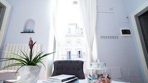 A room with a view of buildings through a window, a potted plant, a chair, and a table with books and a jar of candies. - Le Erbe Guest House (Viterbo)