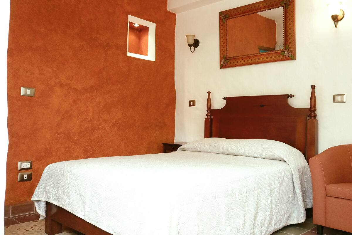 A bedroom with a wooden bed, a white bedspread, a brown chair, a wall-mounted mirror, and a light fixture.