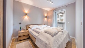 Un dormitorio moderno con cabecera de madera, dos lámparas de noche, una cama grande con ropa blanca y vistas a un balcón a través de una puerta corrediza.