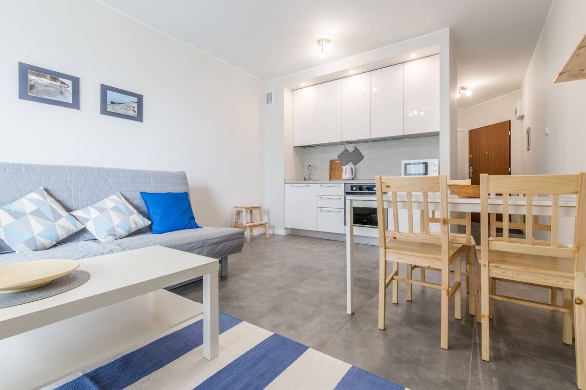 A modern living room with a grey sofa, a white coffee table, and a blue and white striped rug. There is a kitchen area with white cabinets and a wooden dining table with chairs.