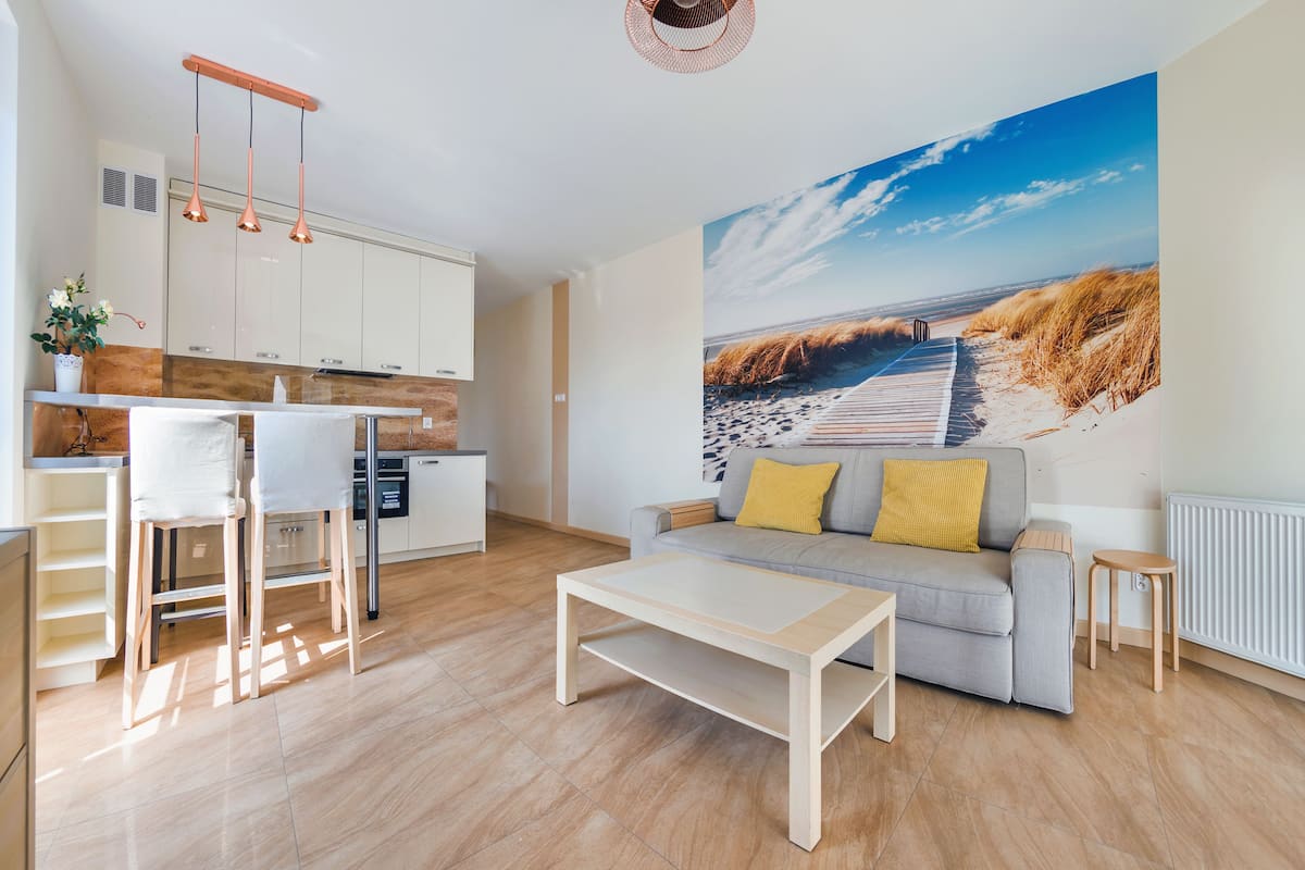 A modern living room with a sofa, coffee table, and a view of a beach through a large window.
