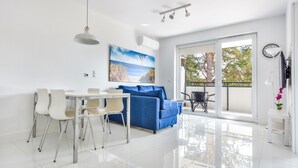 Un salón moderno con una mesa de comedor blanca, un sofá azul y un ventanal con vistas a árboles y un edificio.
