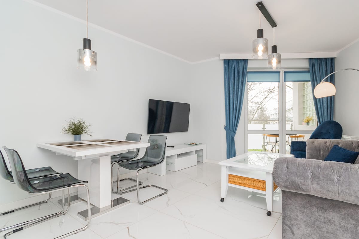 A modern living room with a sofa, a dining table with chairs, a TV, and a large window with curtains.