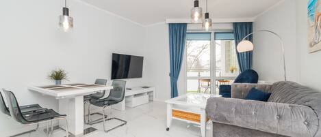 A modern living room with a sofa, a dining table with chairs, a TV, and a large window with curtains.