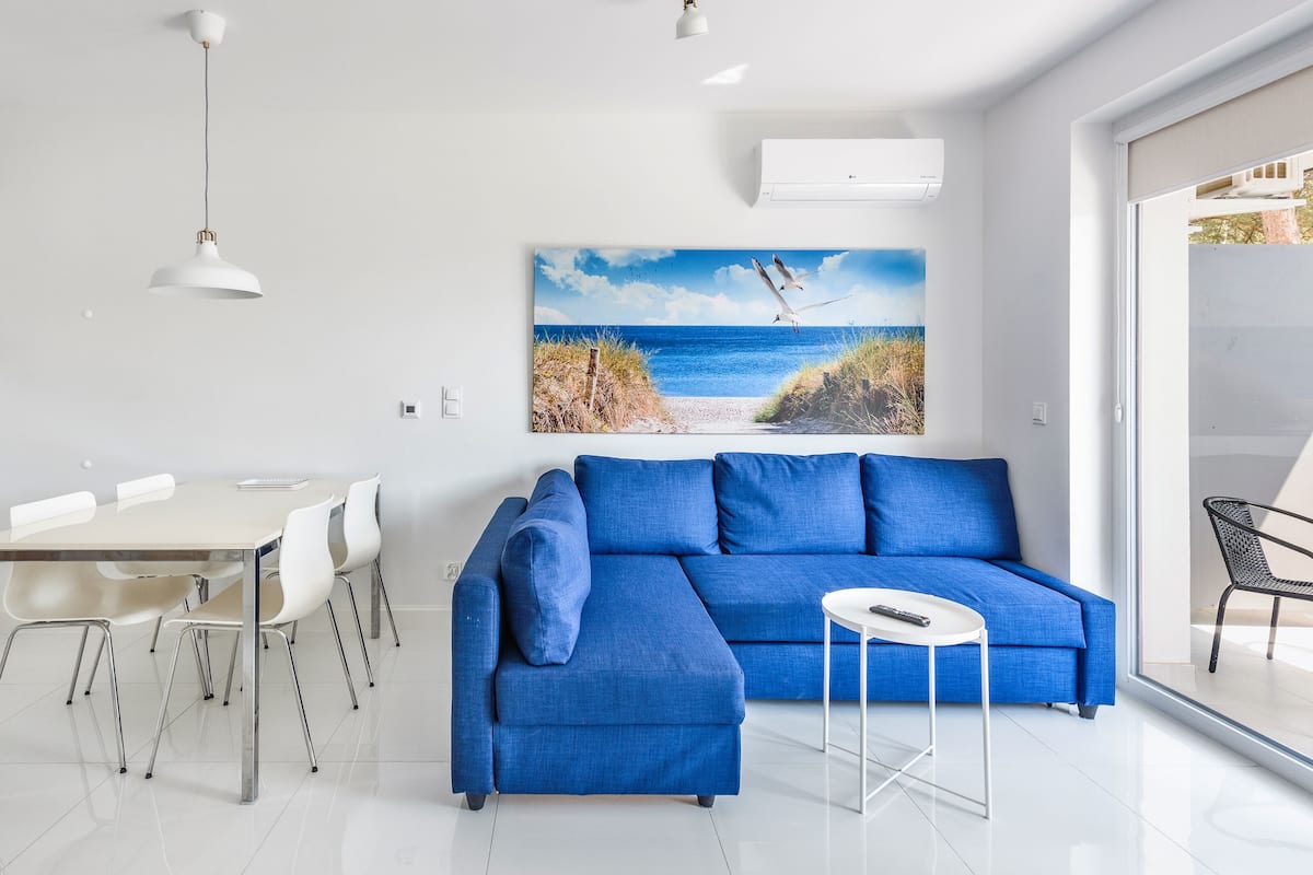 A modern living room with a blue sofa, a dining table with chairs, and a large beach-themed painting on the wall.