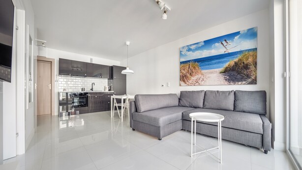 Un espacio habitable moderno con un sofá seccional gris, una mesita blanca y una cocina con salpicadero blanco y armarios oscuros.