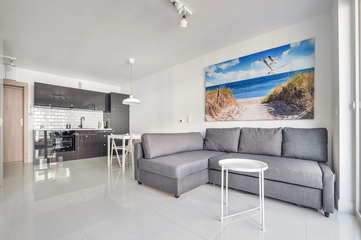 A modern living space with a grey sectional sofa, a small white table, and a kitchen area with a white backsplash and dark cabinets.