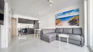 A modern living space with a grey sectional sofa, a small white table, and a kitchen area with a white backsplash and dark cabinets. - Sun & Snow Bursztynowe Osiedle (Stegna)