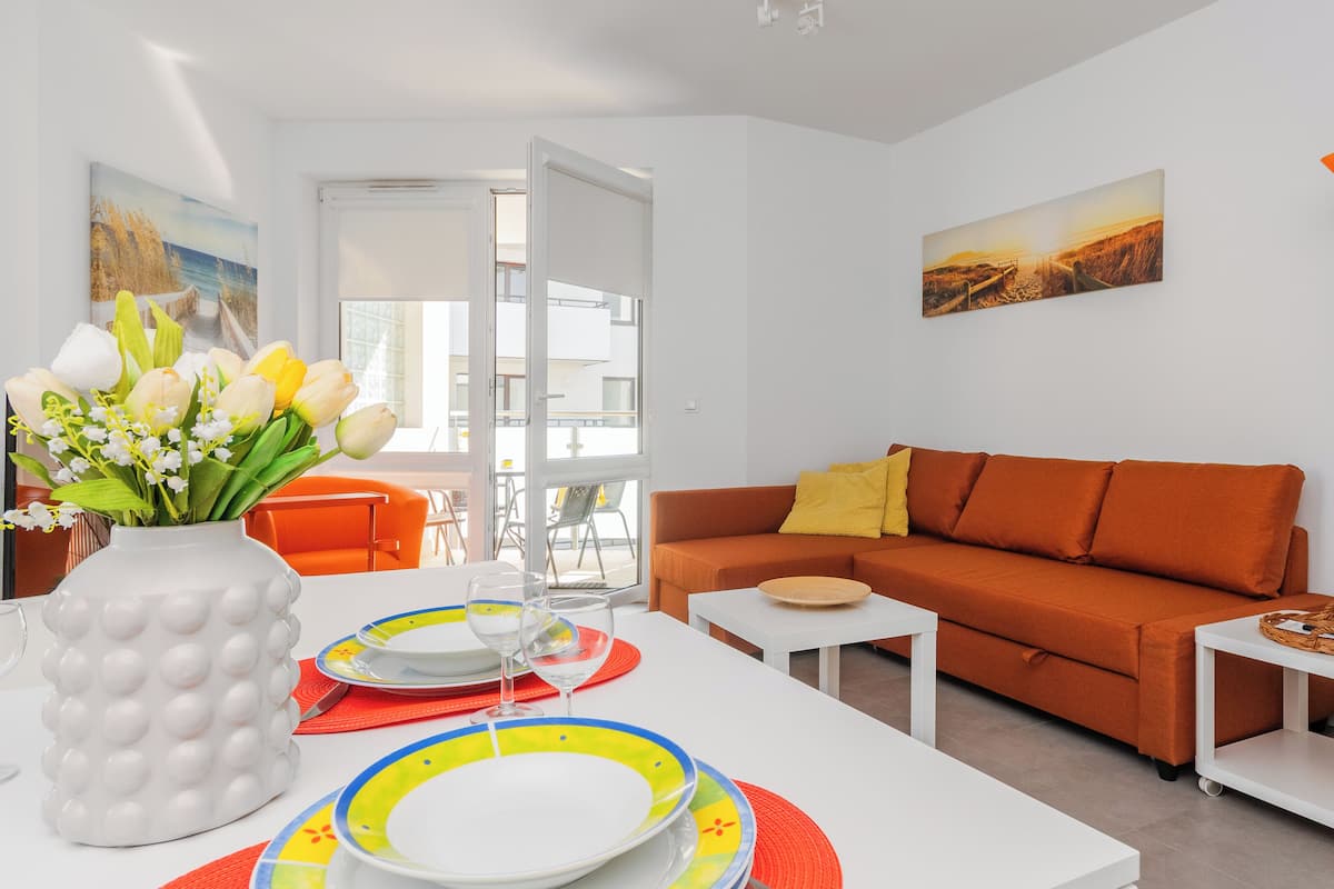 A modern living room with a white sofa, orange cushions, a dining table with colorful plates, a vase with flowers, and a painting on the wall.
