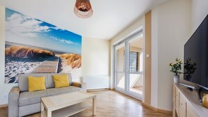 Un salón moderno con un gran mural de playa, un sofá con cojines amarillos, una mesa de centro, un televisor sobre un mueble de madera y un ventanal amplio.