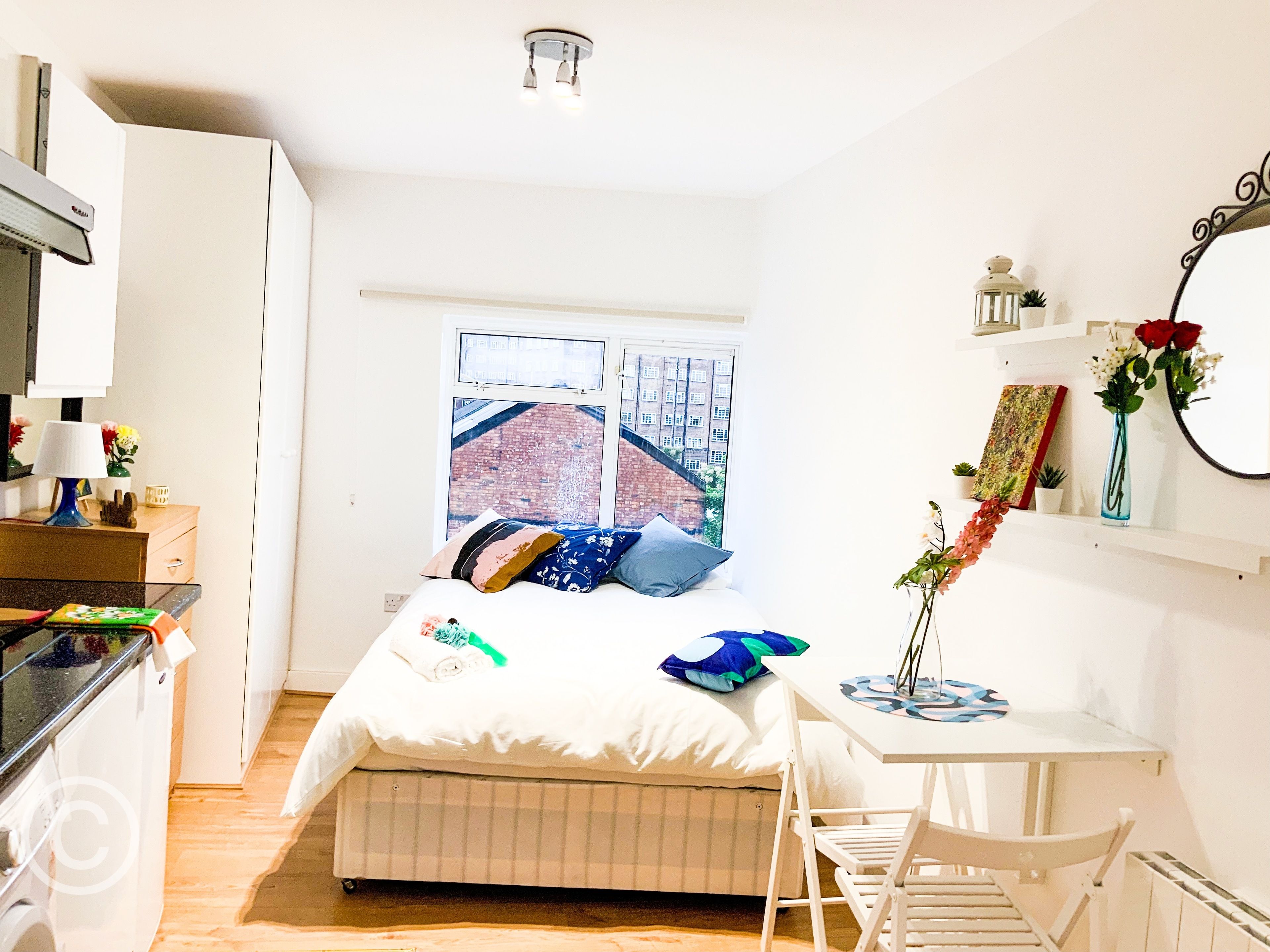 A compact bedroom with a bed, bedside table, and a window with a view of a brick building.