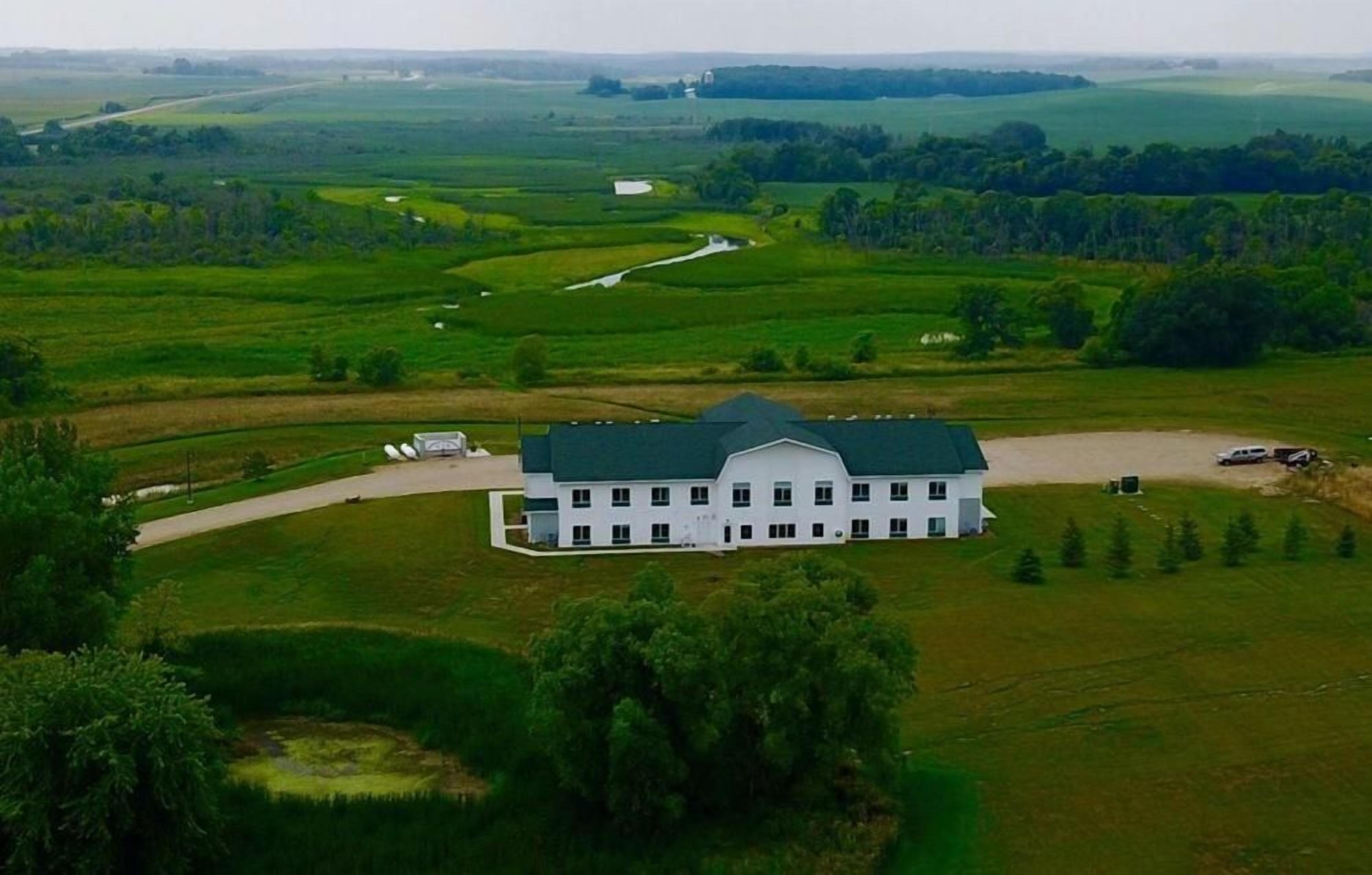 Un grand bâtiment blanc surmonté d’un toit vert, entouré de champs verdoyants et d’arbres.