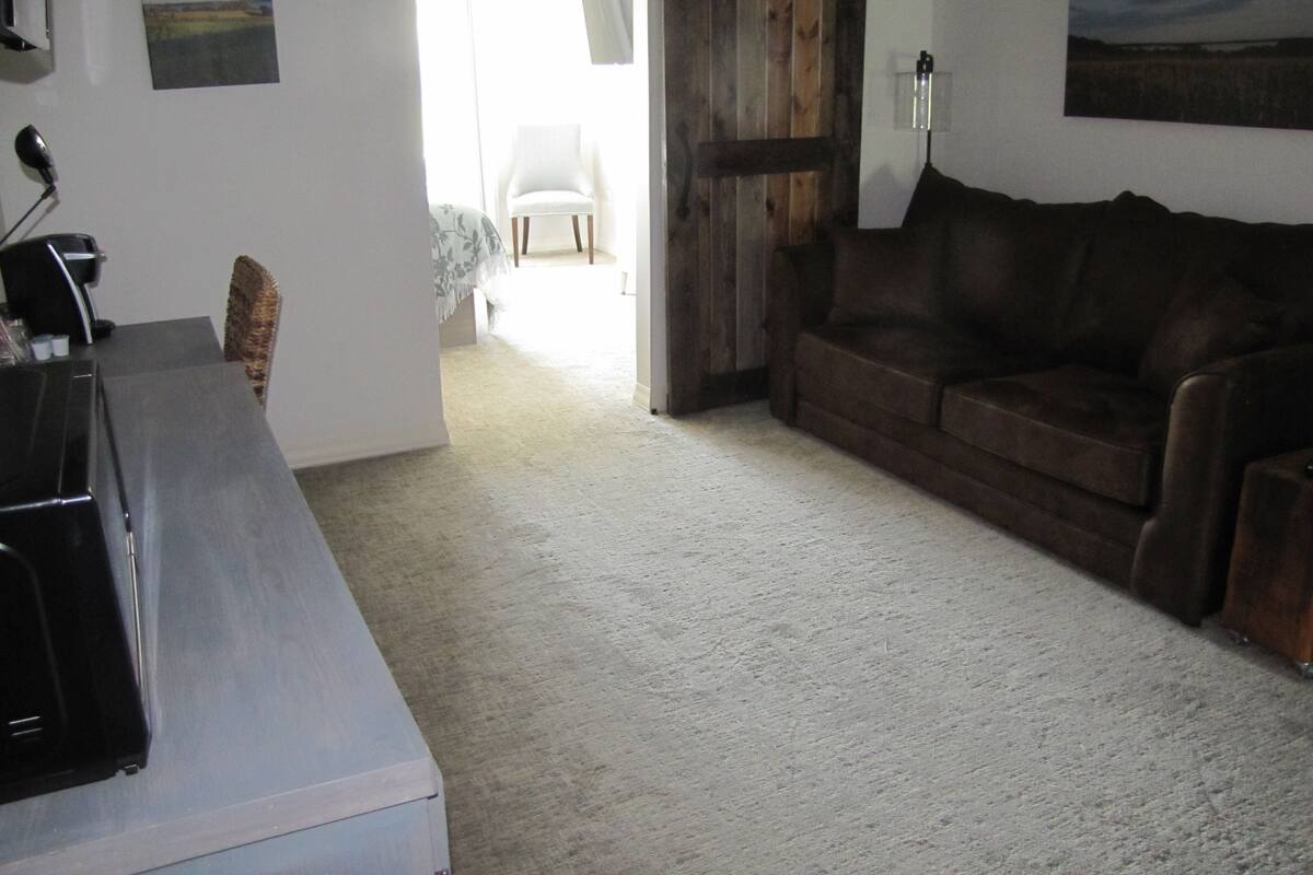 Une pièce avec une porte en bois, une chaise blanche, un canapé marron et un bureau avec une lampe.