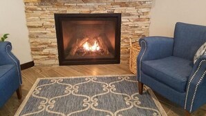 A stone fireplace with a fire, flanked by blue upholstered chairs and a patterned rug. - Lakes Inn at Dunvilla (Pelican Rapids)