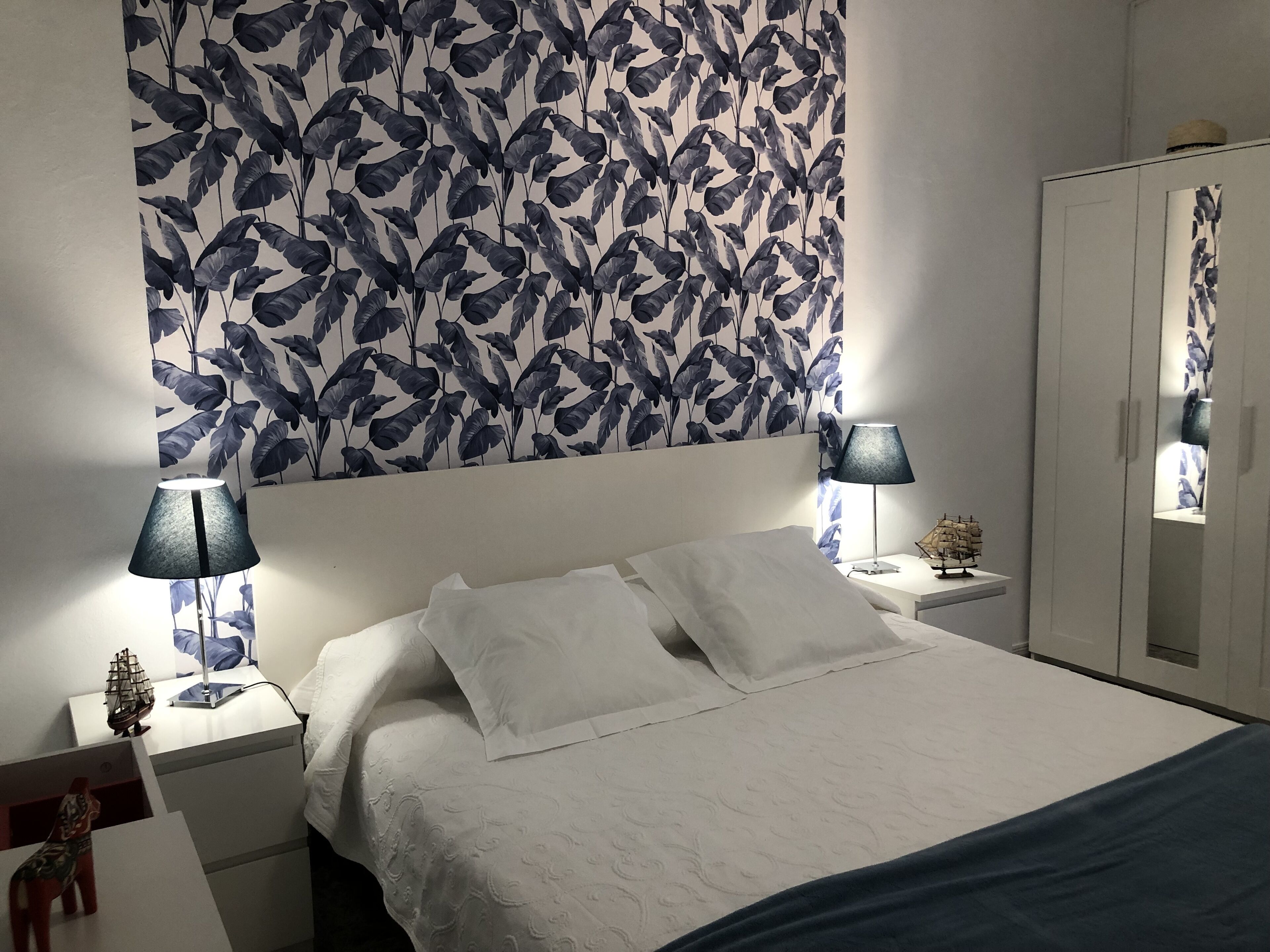 Uma cama bem arrumada com lençóis brancos, duas luminárias de cabeceira, um criado-mudo com um modelo de navio e um guarda-roupa com espelho.