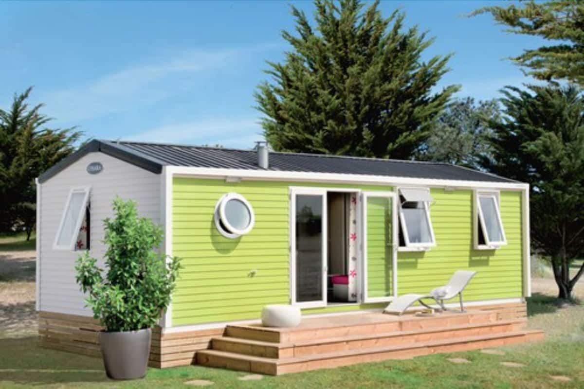 Een kleine, gelijkvloerse stacaravan met een groene buitenkant, een veranda met een bankje en een bloempot.