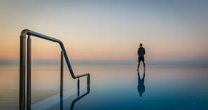 Une personne se tient au bord d’une piscine à débordement dont la surface réfléchit l’environnement, avec une rambarde en métal, et surplombe une étendue d’eau calme au coucher du soleil.